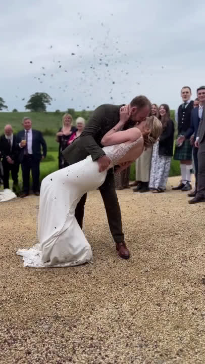 A couple walks through a colourful confetti line and shares a romantic dip kiss for the crowd and camera at their rustic outdoor wedding in Northumberland, UK. This beautiful moment captures the love and charm of a nature-inspired celebration