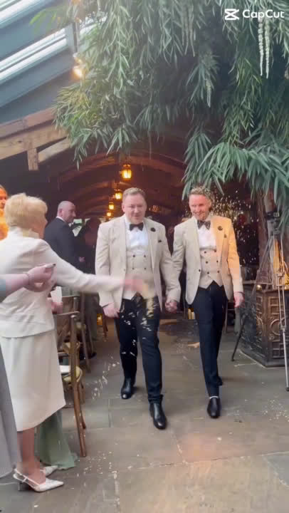 Two grooms walk up the aisle in beige linen suits as confetti is thrown, celebrating their classy New Year's Eve wedding in Northumberland, UK. This joyful moment captures the elegance of their special day, offering modern wedding inspiration